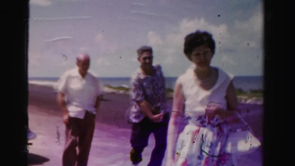 1955 Dade County 8mm frame of three adults walking along a coastal road, with the ocean horizon, clouds, and part of a parked white car visible.