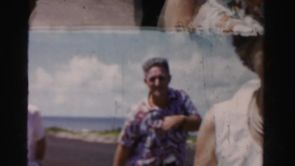 Soft 8mm frame from the 1955 Dade County clip with a man in a patterned shirt near the coastal road and a woman partly cropped in the foreground.