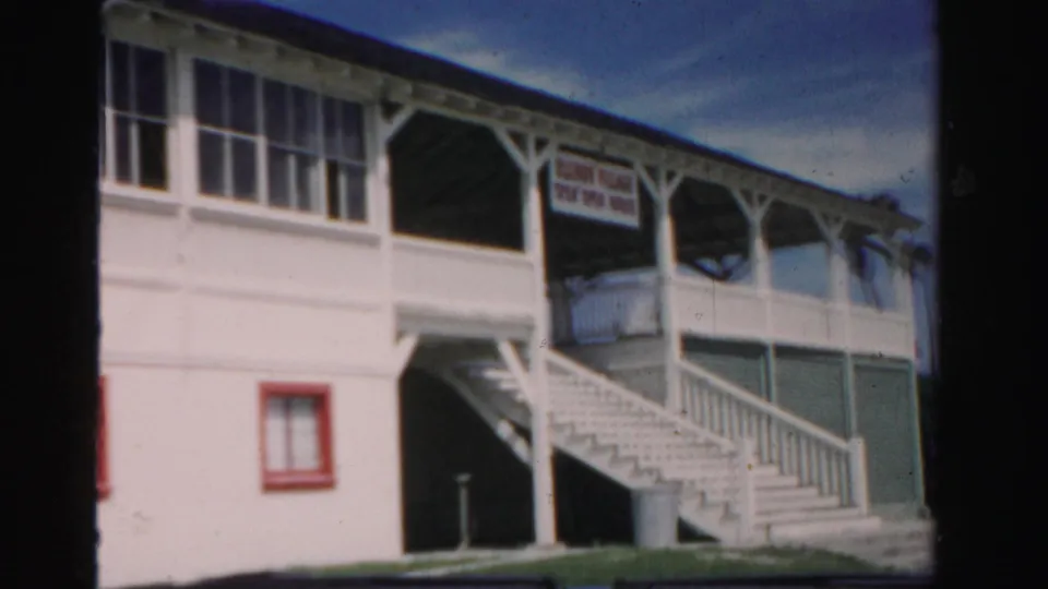 1955 Dade County 8mm frame showing a white two-story roadside building with exterior stairs, red-trimmed openings, and an unreadable sign.