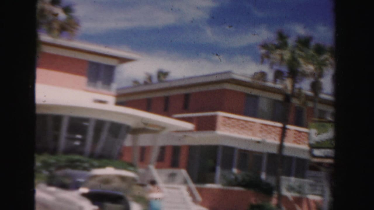 Pink-and-white low-rise building with palm trees seen from a moving car.