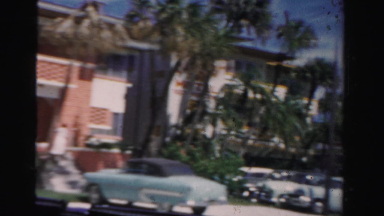 Light blue convertible parked near a palm-lined low-rise building.
