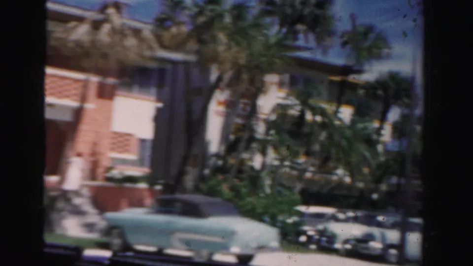 1955 Dade County street view with a light blue convertible, nearby parked cars, palms, and a pink-and-white building.