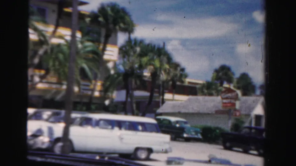 1955 Dade County frame with a white station wagon, motel sign, palms, and low roadside buildings.
