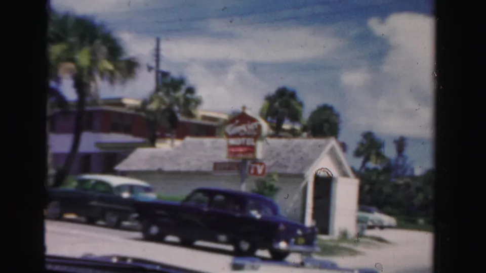 1955 Dade County street frame showing a dark sedan, motel sign, small white roadside structure, and palm trees.