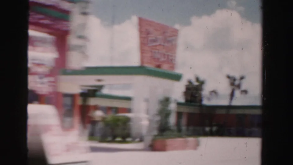 1955 Dade County roadside view showing a white building with green trim and an unreadable red sign, seen from a moving camera.