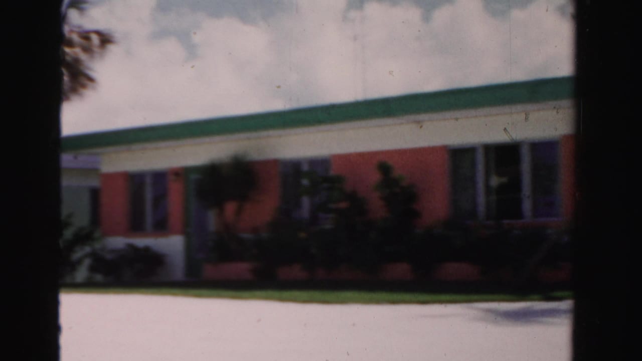 Low coral and white building with green roof trim, lawn, shrubs, and palm shadows.