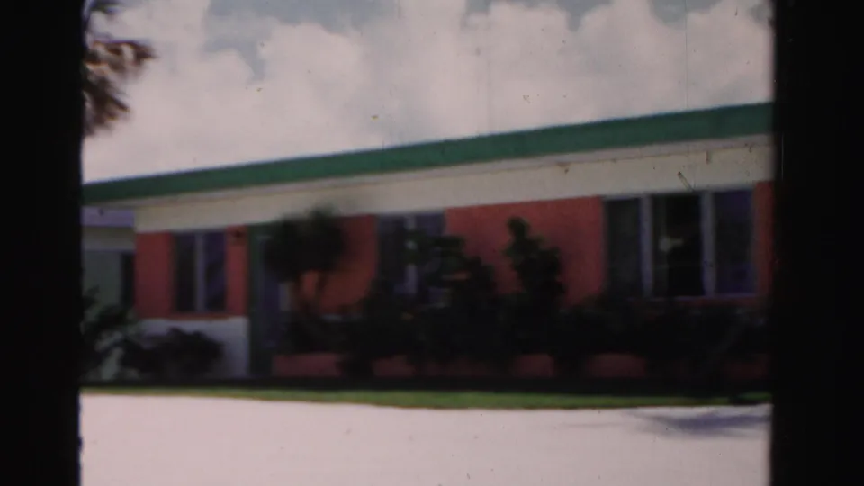 1955 Dade County exterior of a low coral-and-white building with green roof trim, lawn, shrubs, and bright sun.