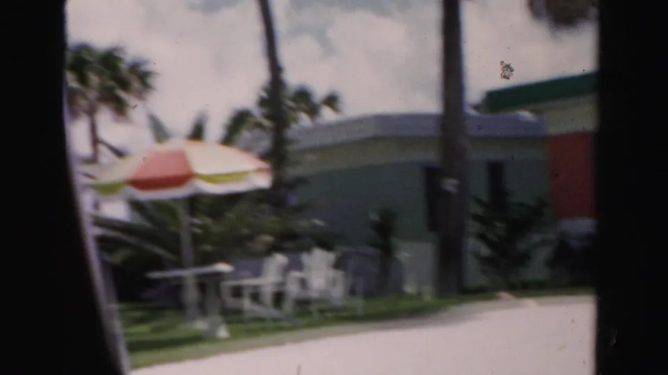 1955 Dade County lawn scene with patio furniture, a striped umbrella, palms, and a low green-and-coral building.