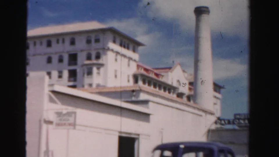 1955 Dade County view of a large white structure, tall chimney, low foreground building, and dark car under a bright sky.