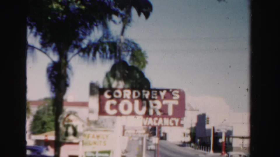 1955 Dade County 8mm frame showing Cordrey's Court signage, palm trees, low roadside buildings, and a parked car.