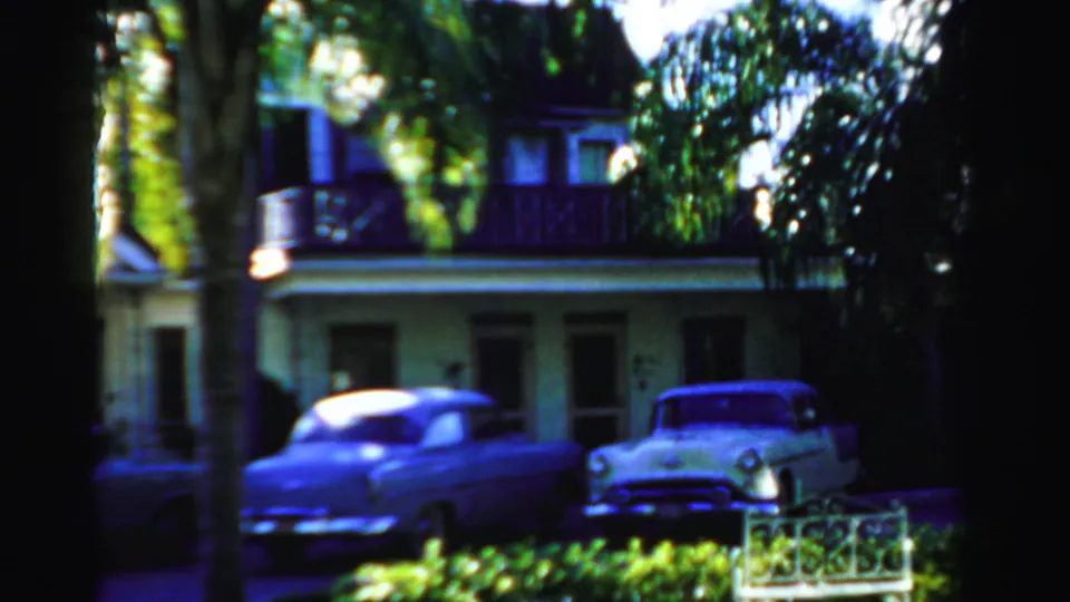 1955 Dade County frame showing parked mid-century cars, a shaded porch-front building, palms, and low curbside planting.