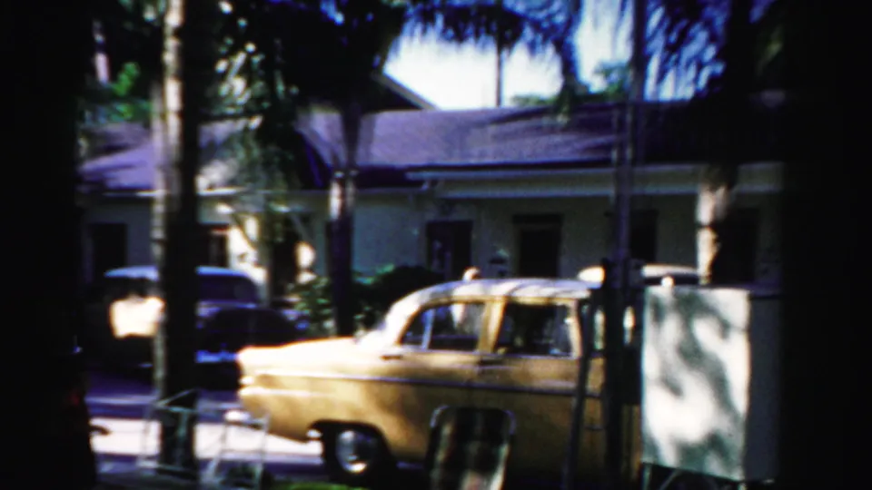 1955 Dade County home-movie frame showing a yellow parked car, palm trunks, and a sunlit stucco residential building.