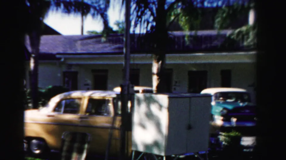 1955 Dade County street view with yellow and turquoise parked cars outside a low stucco building under palm shade.