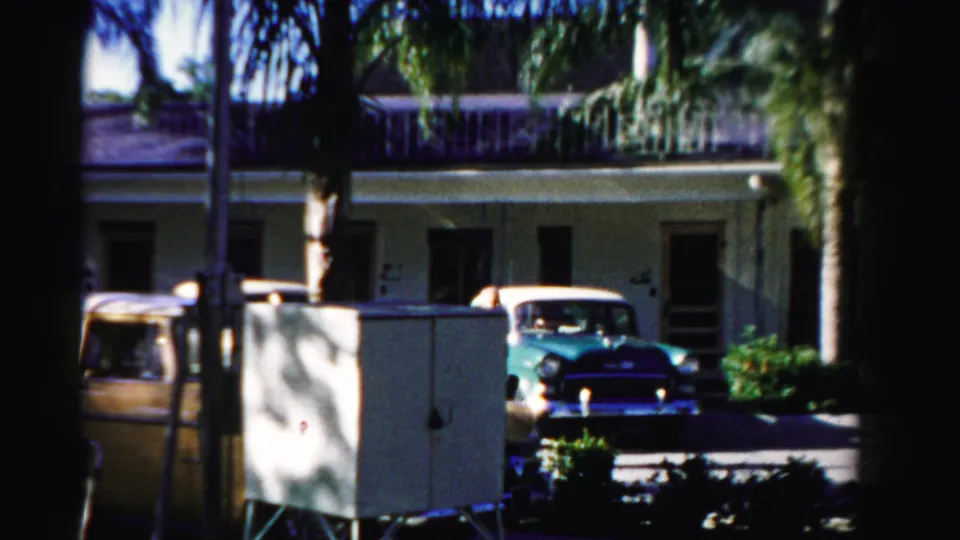 1955 Dade County frame with a turquoise parked car, partial yellow car, palm trees, and a stucco residential exterior.