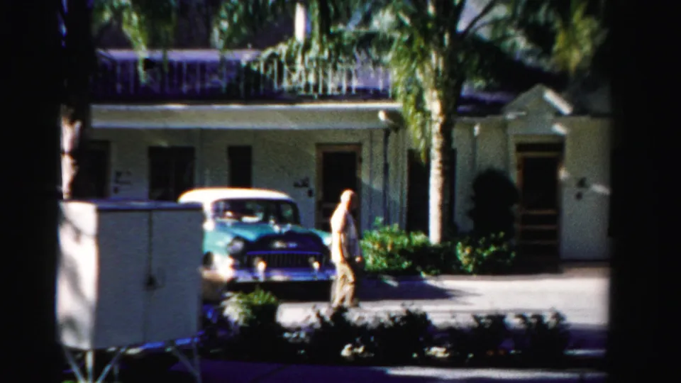 1955 Dade County home-movie frame with a man walking past a turquoise car near a palm-lined stucco building.
