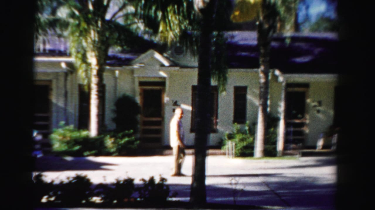 Man in light clothing walking past a low building and palm trees.