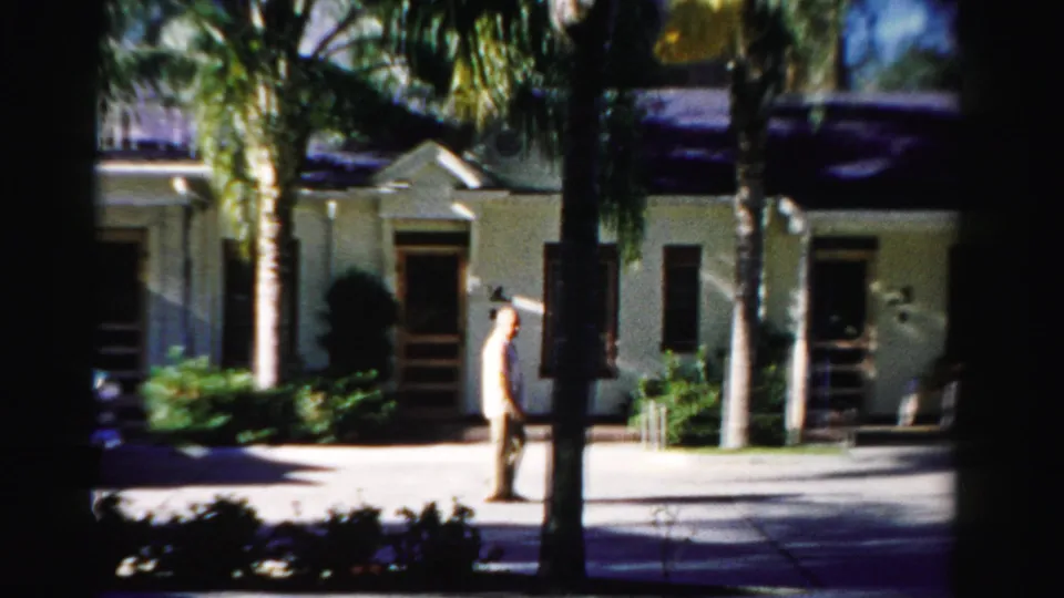 The man continues along the sidewalk in a 1955 Dade County street view, with palm trunks and a cream building behind him.