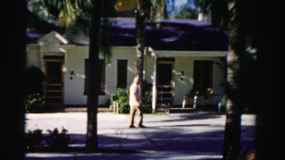 A mid-1950s Florida home-movie view shows the man farther along the sidewalk beside palms, windows, and a shaded doorway.