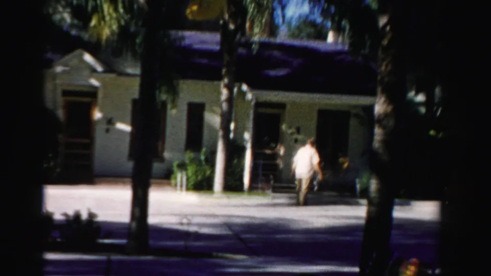 At the end of the clip, the man approaches the right side of the Dade County building exterior, partly framed by palm trunks and shadows.