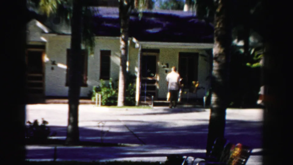 The figure nears the doorway of a modest Dade County house, seen through dark palm shadows on 8mm film.