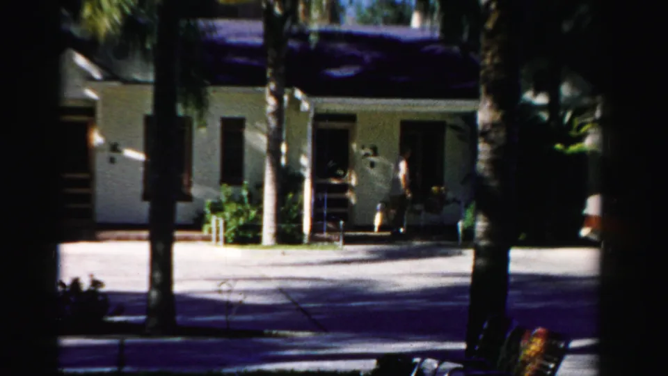A person stands close to the front doorway of the Florida house, with palms and bright driveway light in the frame.