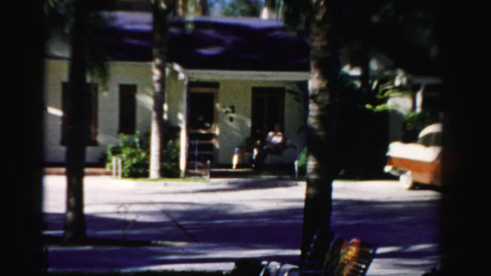 In 1955 Dade County home-movie footage, a two-tone sedan approaches a driveway beside palm trees and a low house.