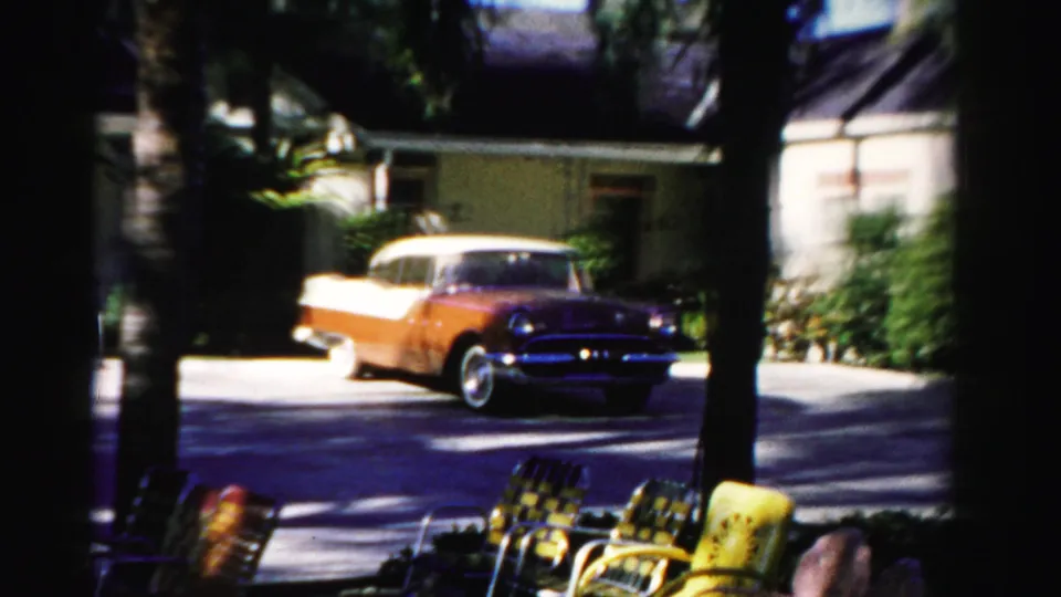 The sedan continues through a residential frame with patio chairs in the foreground and a palm-lined house behind it.