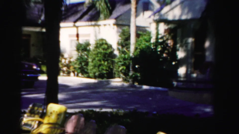 The final frame holds on the residential driveway area, with palms, shrubs, patio chairs, and the car partly leaving view.