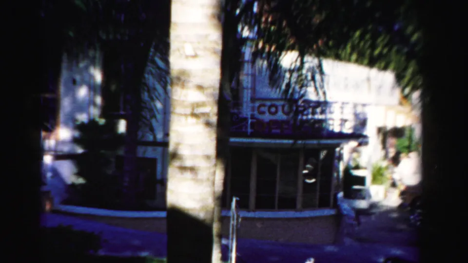 1955 Dade County frame showing a palm trunk, shaded entrance, parked car area, and a partially readable building sign.