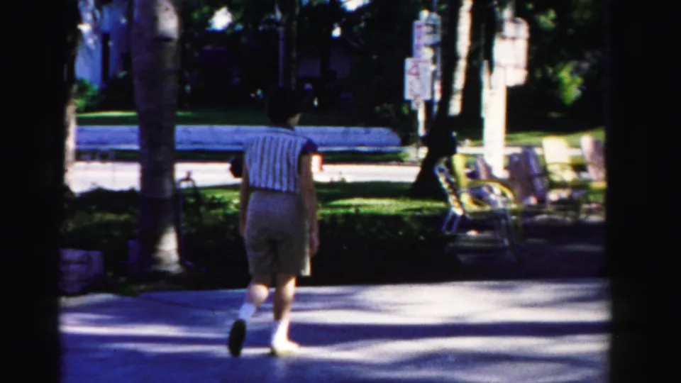 1955 Dade County 8mm frame of a woman in tan shorts walking away near palms, benches, greenery, and a street sign.