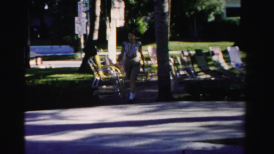In 1955 Dade County, Florida footage, a woman walks along a shaded resort path beside palm trees, lawn chairs, and grass.