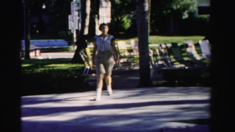A woman in a striped top and shorts walks toward the camera on a sun-dappled palm-lined walkway in Dade County, Florida.