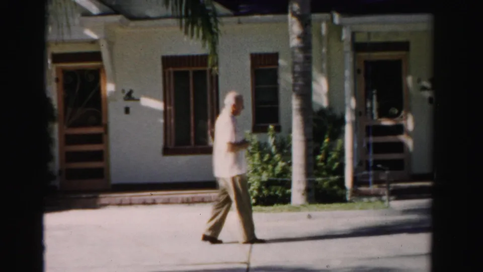 An older man in a light shirt and khaki trousers walks past a white stucco building with palm trunks in the foreground.