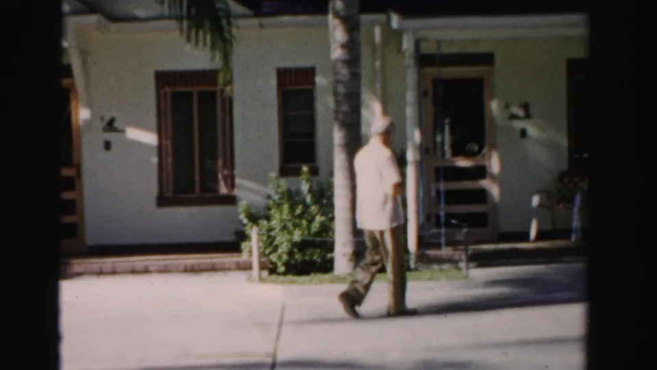 An older man walks across the frame beside palm trees and a white stucco building, with deep shade around the entrance area.