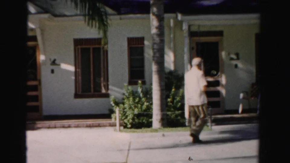 1955 Dade County home-movie frame showing a man near a sidewalk, palm trunks, shrubs, and a white stucco house entrance.