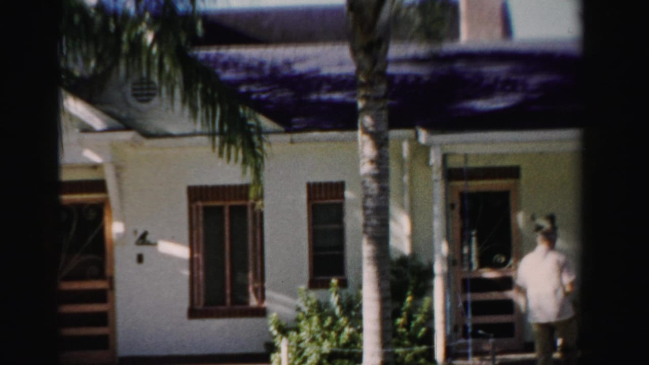 1955 Dade County exterior frame with a man by a shaded doorway, palm trunks, windows, and a white stucco residential facade.