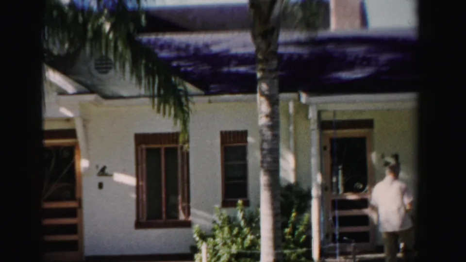 1955 Dade County exterior frame with a man by a shaded doorway, palm trunks, windows, and a white stucco residential facade.