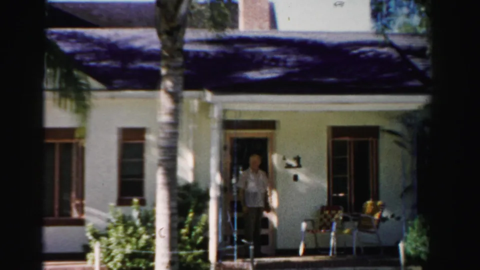 1955 Dade County home-movie frame of a white house porch with palm tree, chairs, windows, and a man in the doorway.