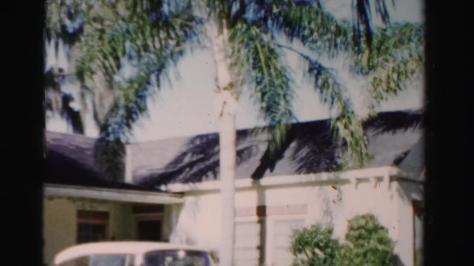 The downward pan reveals more of the 1955 Dade County house, including shaded windows, shrubs, palms, and part of a light-colored car.