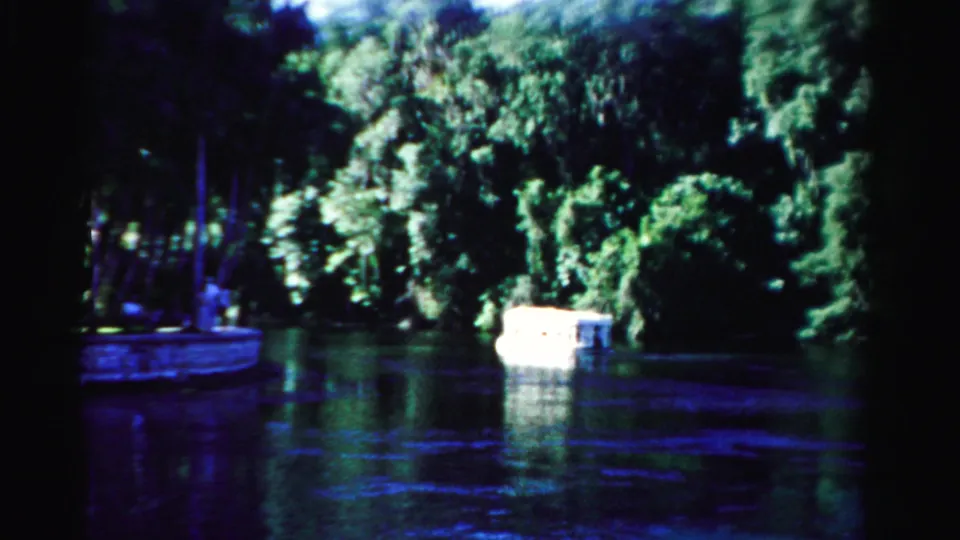 1955 Dade County home-movie frame showing a small white boat moving along dark water with heavy green shoreline trees behind it.