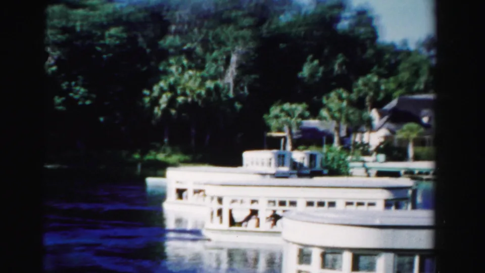 A white passenger boat passes across calm water near palm trees and waterfront houses in 1955 Dade County, Florida footage.