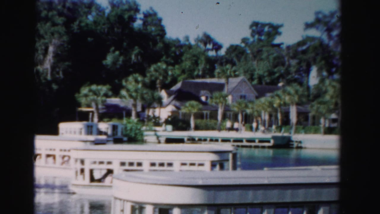 The boat moves farther across the frame, with a dock, palms, and large waterfront houses visible on the Dade County shoreline.