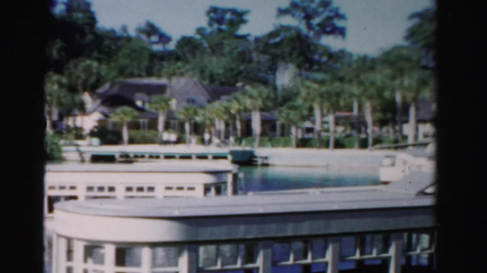 The passing boat fills the lower frame while a palm-lined waterfront and house remain visible in the 1955 Florida home-movie shot.