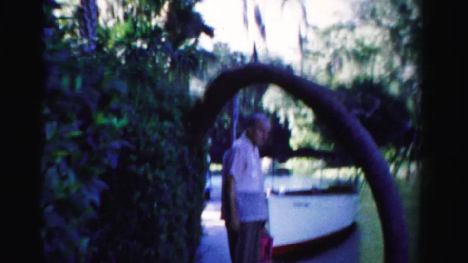 1955 Dade County frame showing an older man in light clothing beside a white boat, lake, palms, and arched tree limb.