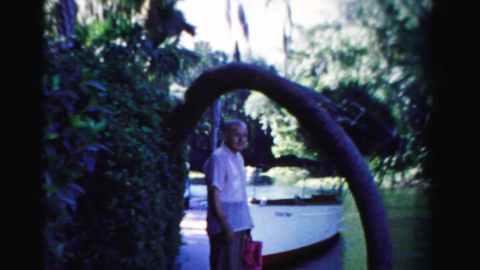 1955 Dade County home-movie frame showing a man on a shaded path beside a curved tree limb, docked white boat, water, and dense greenery.