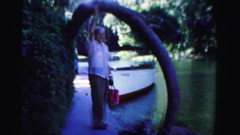 1955 Dade County home-movie frame showing a man under a curved tree arch, holding a red bag beside a docked boat and shaded waterway.