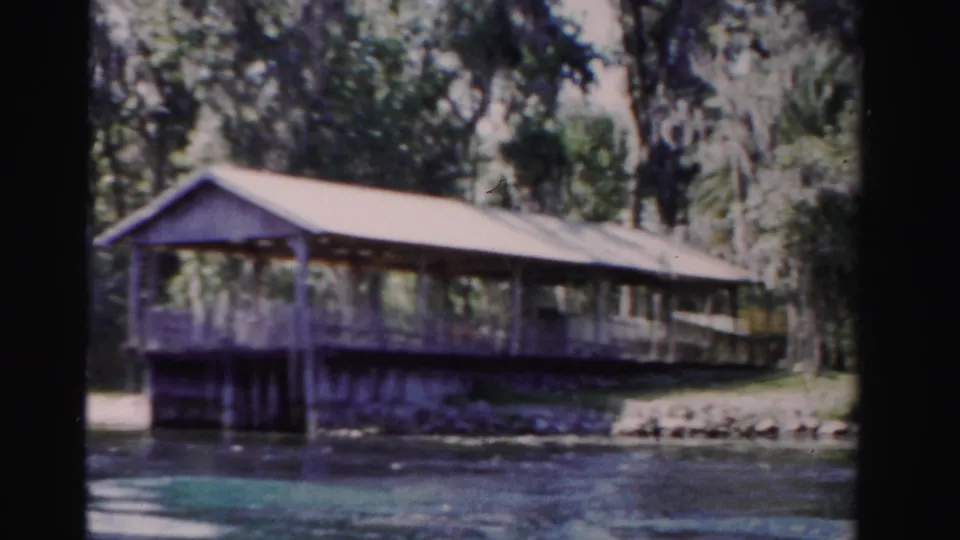 Soft 8mm frame of a covered lakeside deck in Dade County, Florida, with calm water and shaded trees in the background.
