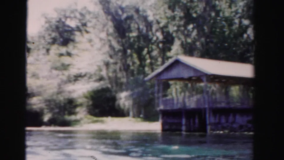 End of the pan shows the covered deck at frame edge, calm lake water, sunlit bank, and dense trees in 1955 Dade County footage.