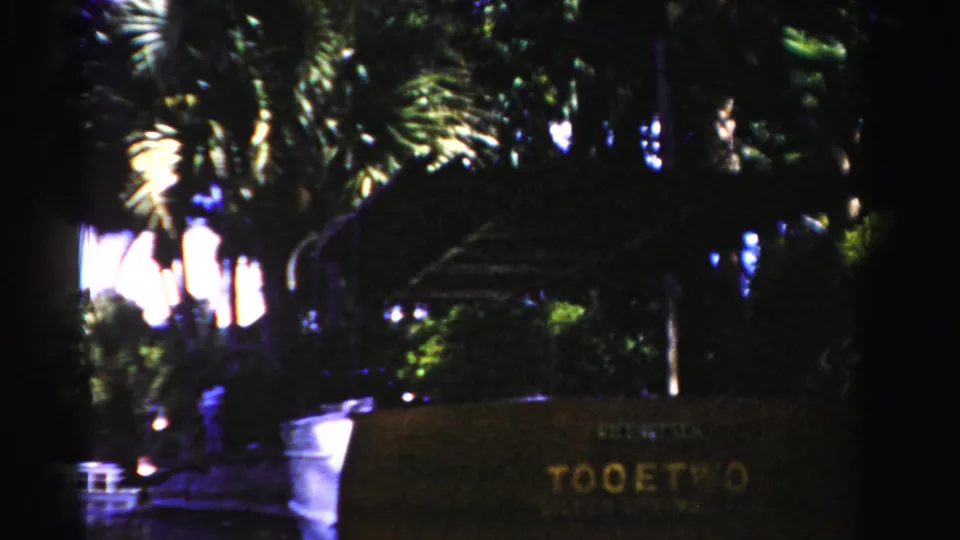 1955 Dade County 8mm frame showing a dim waterfront area with palm trees, a white boat, dark water, and a brown sign reading TOOETWO.