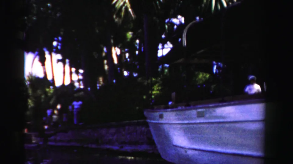 1955 Dade County frame showing the white boat, palm trees, dock area, and a faint person in the dim background at right.